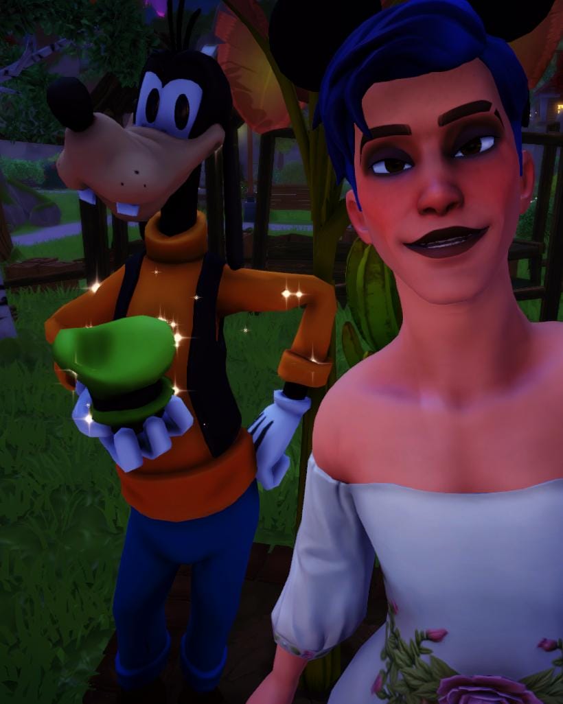 A selfie of the player character and Goofy. The player has a masculine body frame, blue hair, and is wearing a white gown with floral details on the waist. They are also wearing black eyeshadow and black lipstick.