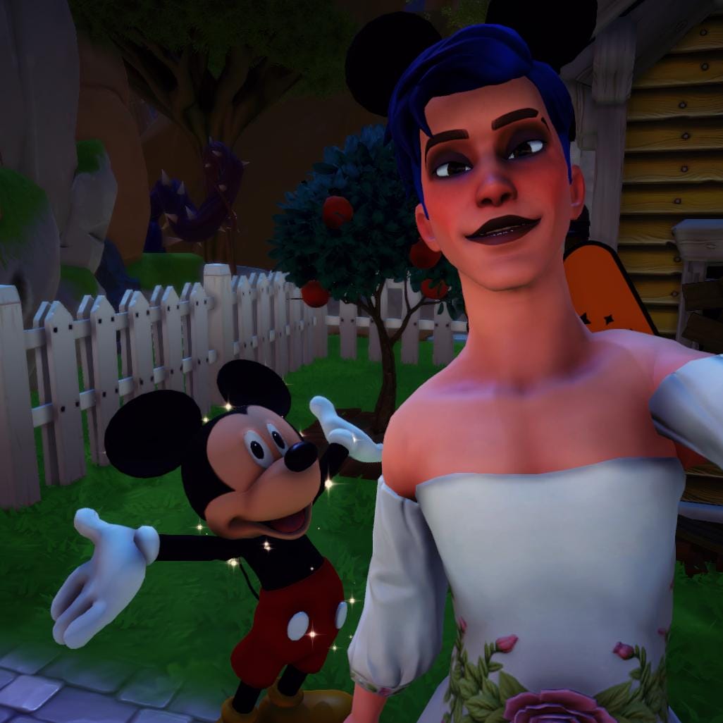 A selfie of the player character and Mickey Mouse. The player has a masculine body frame, blue hair, and is wearing a white gown with floral details on the waist. They are also wearing black eyeshadow and black lipstick.
